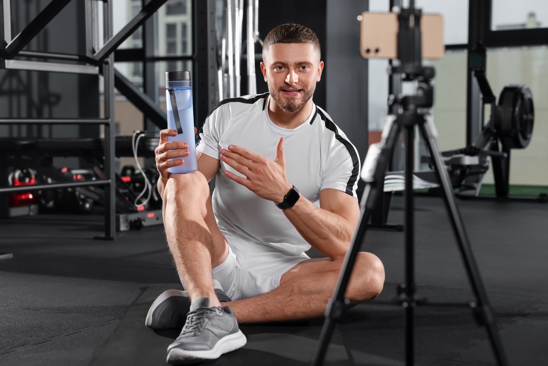 Man with bottle of water streaming online training on phone at gym. Fitness coach